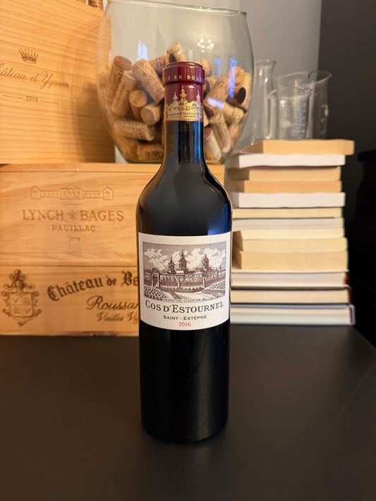 Bottle of Chateau Cos d'Estournel 2016 wine with wooden boxes and books in the background.