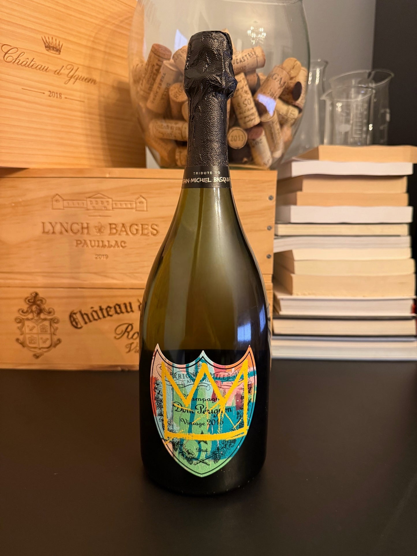 Bottle of Dom Pérignon Vintage 2015: Basquiat Special Edition with a wooden box and books in the background.