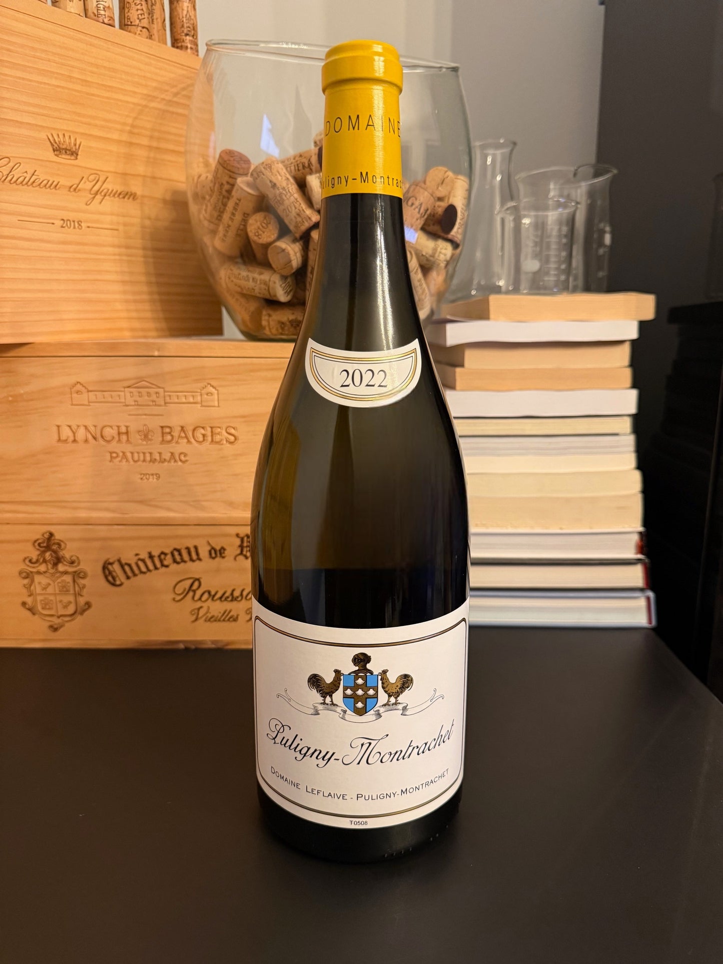 Wine bottle with a Domaine Leflaive Puligny-Montrachet 2022 MAGNUM in front of wooden wine boxes and books.