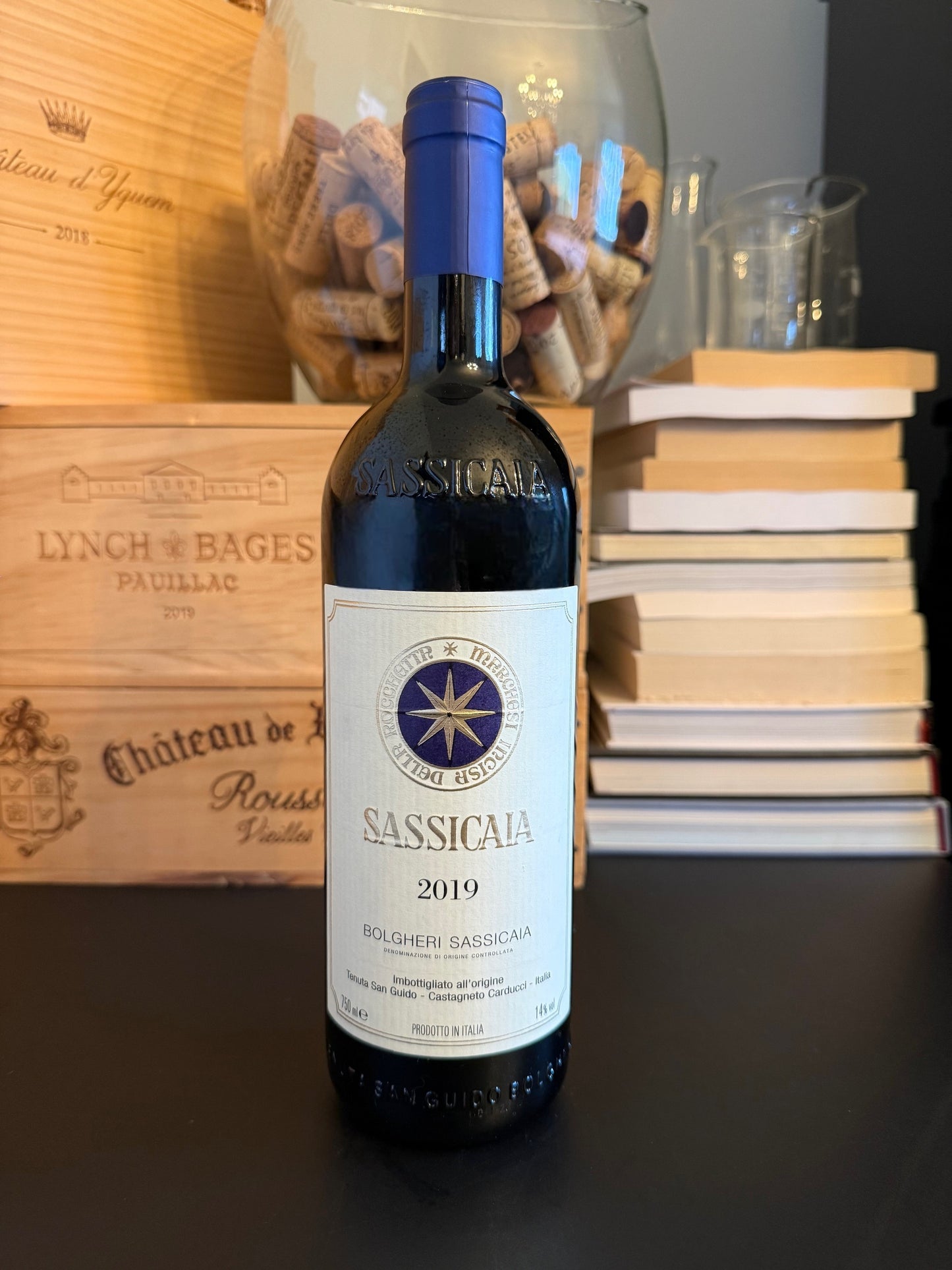 Bottle of Sassicaia 2019 wine with a wooden box and books in the background.